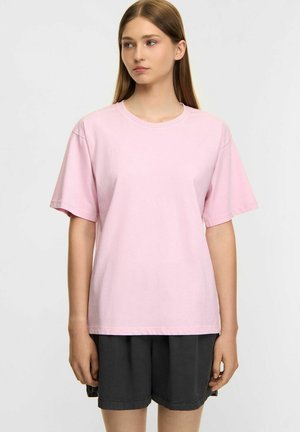 Young woman with straight light brown hair wearing plain light pink oversized t-shirt and black shorts, standing against a plain white background.