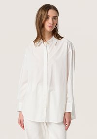 Cămașă albă cu butoni, fabricată dintr-un material ușor, având un guler clasic, mâneci lungi și o croială lejeră cu un tiv curbat.