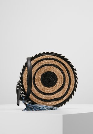 Runde geflochtene Strohtasche zum Umhängen mit schwarzen konzentrischen Kreisen, schwarzem Lederriemen und blau-weißem Quaste auf weißem Sockel.