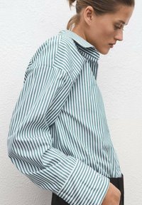 Striped long-sleeve shirt in green and white, featuring a structured collar, relaxed fit, and wide cuffs with vertical lines. Soft fabric texture.