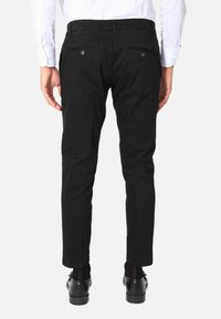 Pantaloni neri a vestibilità slim, con tasche posteriori e texture liscia, indossati con scarpe nere e una camicia bianca.