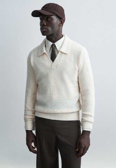 Cream textured knit sweater with a v-neck, paired with a white collared shirt and a patterned brown tie, topped with a brown cap.