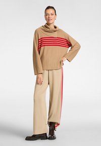 Maglione oversize beige con strisce orizzontali rosse, dotato di collo a camicia. Abbinato a pantaloni beige a gamba larga con strisce laterali rosse e stivali neri.