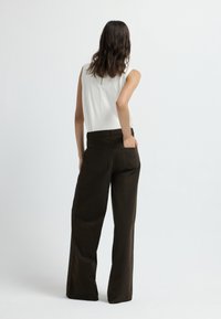 Pantaloni larghi marroni con bottoni laterali, tessuto strutturato e tasche posteriori. Indossati con un top bianco senza maniche, vista da dietro.