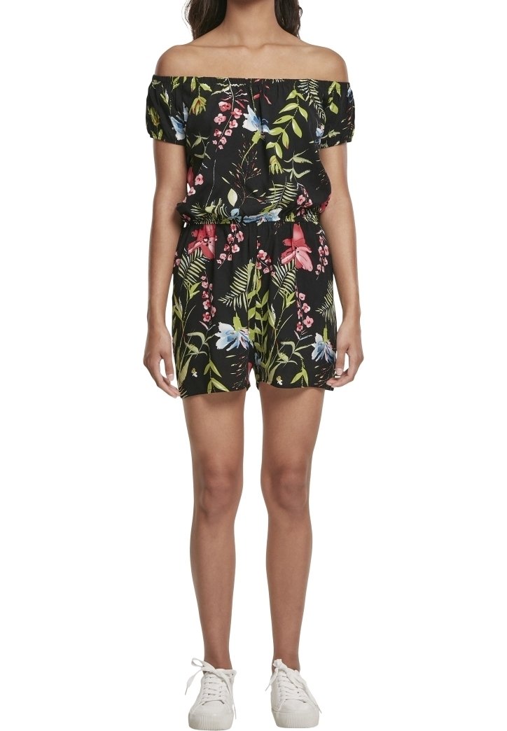 Femme portant une combinaison noire à fleurs épaules dénudées avec des feuilles vertes, rouges et bleues, associée à des baskets blanches.