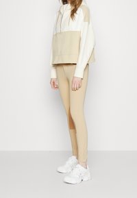 Beige zkrácená mikina s bílým vrškem, klokanní kapsou a šňůrkou. Béžové legíny se bočními panely a bílé sportovní boty.