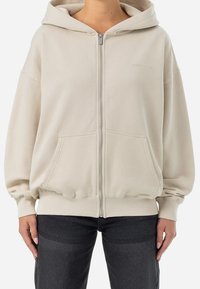 Beige zip-up hoodie i mjukt material; har en huva, oversize passform och framfickor med ribbad mudd och nederkant.