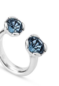 Anillo abierto de plata con dos grandes piedras azules facetadas sujetas por cuatro garras en cada extremo, mostrado sobre un fondo blanco.