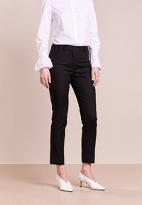 Zwarte, op maat gemaakte broek met een slim fit en een soepele textuur. Voorzien van riemlussen en zijzakken. Gecombineerd met een witte blouse met lange mouwen en witte hakken.