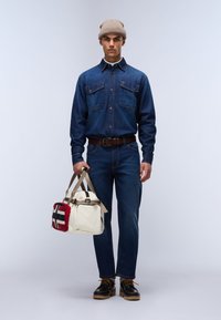Hombre con gorro beige, camisa de mezclilla y jeans, sosteniendo una bolsa color crema con un parche rojo y negro, de pie sobre un fondo claro y liso.