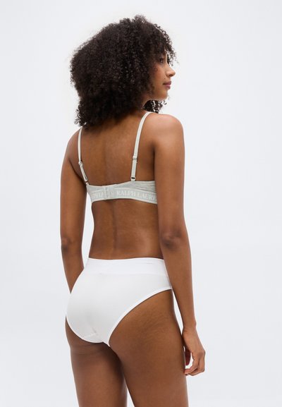 Γυναίκα με σγουρά μαλλιά που φοράει γκρι σουτιέν με το κείμενο "Ralph Lauren" και λευκά εσώρουχα με ψηλή μέση, στέκεται μπροστά σε απλό φωτεινό φόντο.