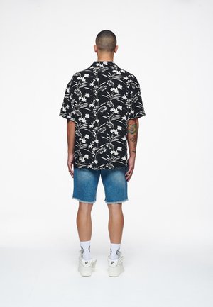 Hombre con cabello corto lleva camisa negra con flores, pantalones cortos de mezclilla, calcetines blancos y zapatillas blancas, de pie de espaldas sobre fondo blanco.