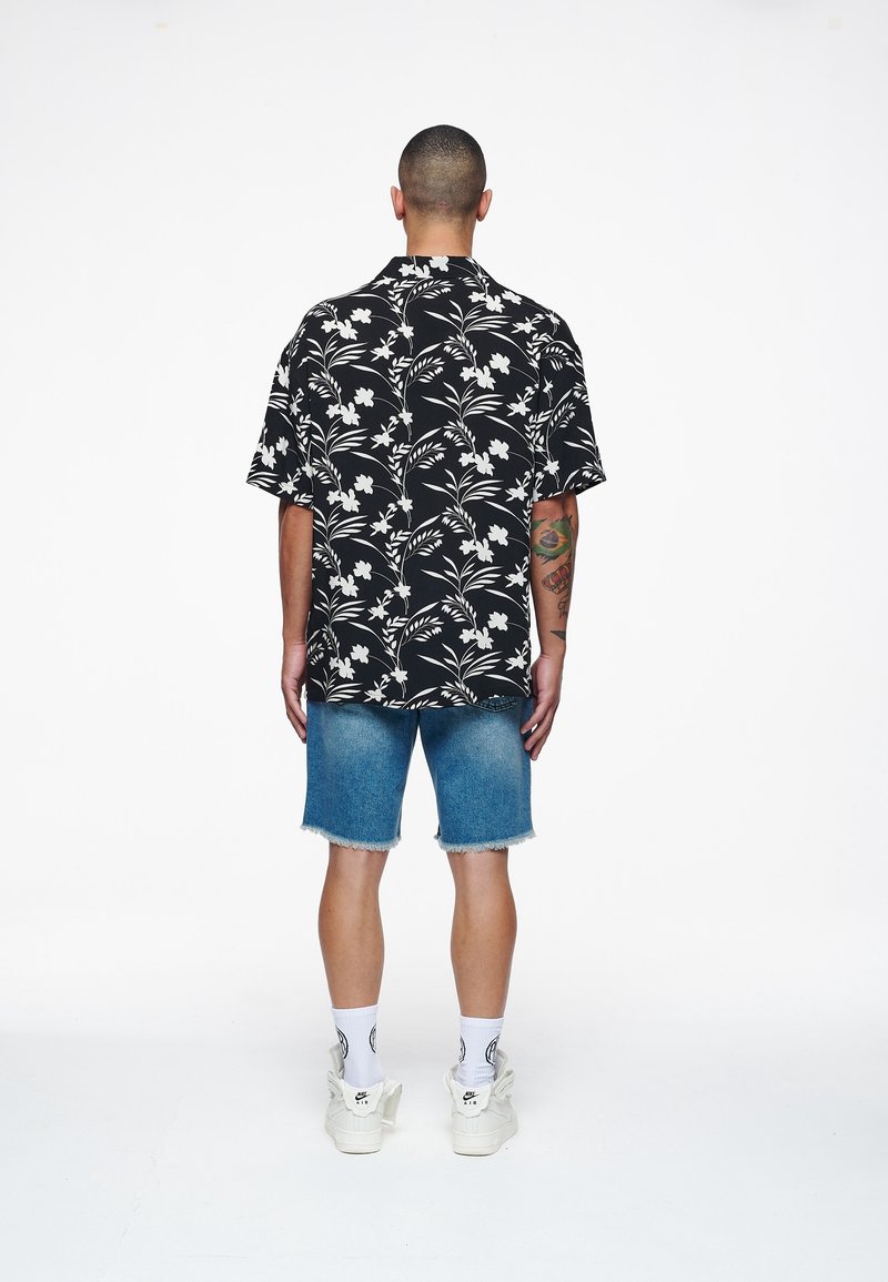Homme aux cheveux courts portant une chemise noire à motifs floraux, un short en denim, des chaussettes blanches et des baskets blanches, debout de dos sur fond blanc.
