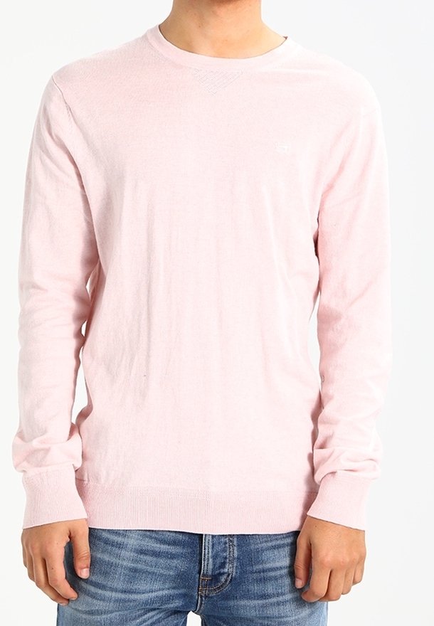Mann in hellrosa langärmligem Pullover und blauen Jeans, der vor einem schlichten weißen Hintergrund steht.