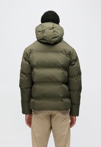 Olívazöld kapucnis pehelydzseki steppelt mintával és sima textúrával, bézs nadrág fölött viselve, hátulnézetből.