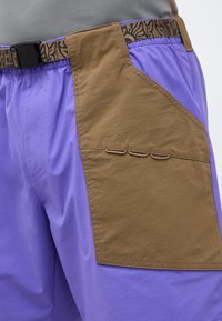 Κοντινή λήψη μοβ σορτς με μεγάλη καφέ πλαϊνή τσέπη και αντίστοιχη ζώνη με σχέδιο και μαύρη πόρπη στη μέση.