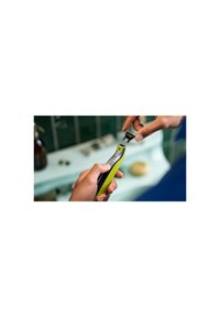 Green and black handheld grooming tool with a textured grip, displaying a removable attachment, set against a blurred background.