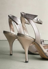 Metallic silver high-heeled sandals feature a knotted bow accent, thin straps, and a curved heel design, showcasing a smooth, reflective texture.