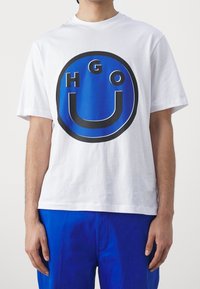 Camiseta de algodón blanca con un gran gráfico circular azul donde aparecen las letras "HGO" en negro y un diseño de cara sonriente.