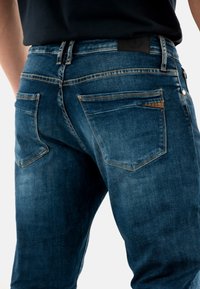 Jeans de mezclilla azul oscuro con corte recto, que presentan costuras en contraste, dos bolsillos traseros y un parche de cuero texturizado en la cintura.