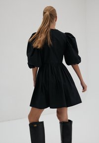 Kobieta z długim kucykiem, ubrana w czarną sukienkę z bufiastymi rękawami i czarnymi kozakami do kolan, stojąca na tle jednolitego białego tła.