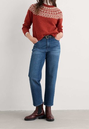 Vrouw draagt een rode trui met patroon, hoge blauwe jeans met zwarte riem en donkerbruine enkellaarzen, handen in de zakken.