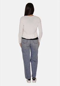 Maglietta bianca a maniche lunghe con dettaglio peplum sul retro, abbinata a jeans azzurri chiari e sneakers bianche con dettagli neri sul tallone.