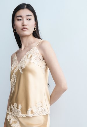 Jeune femme aux longs cheveux noirs portant un caraco en satin beige avec des détails en dentelle florale et des boucles d'oreilles créoles dorées, regardant sur le côté.