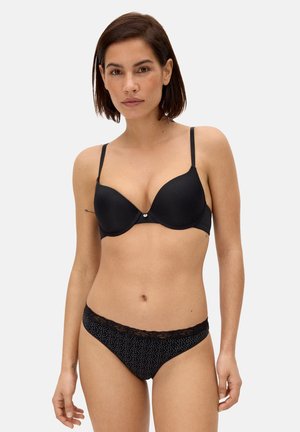 Mujer con cabello corto y oscuro, vistiendo un sostén negro acolchado y bragas negras con lunares y encaje en los bordes, de pie contra un fondo claro y sencillo.