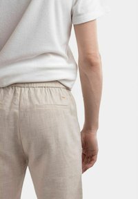 Helle beige Leinenhose mit elastischem Bund, zwei Gesäßtaschen und einem kleinen orangefarbenen Logo. Die Textur ist leicht zerknittert.