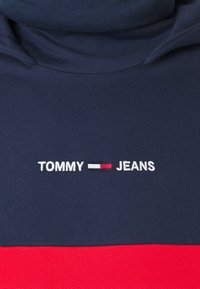 Sweat à capuche bleu marine et rouge avec le texte "TOMMY JEANS" brodé en blanc et un petit logo de drapeau au centre de la poitrine.
