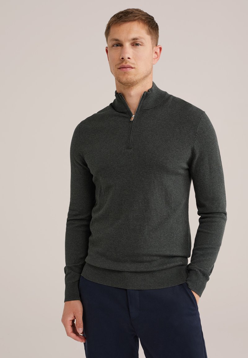 Férfi sötétszürke, negyedzipzáras pulóverben és sötétkék nadrágban, áll egy kézzel a zsebében egy sima bézs háttér előtt.