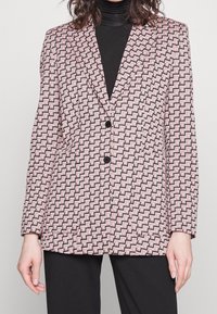Blazer s geometrickým růžovým a černým vzorem, strukturovaným designem, klopami a dvěma černými knoflíky. Materiál se zdá být hladký s texturou.
