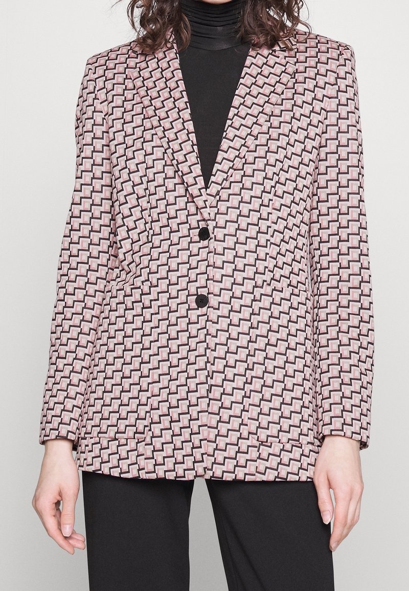 Blazer s geometrickým růžovým a černým vzorem, strukturovaným designem, klopami a dvěma černými knoflíky. Materiál se zdá být hladký s texturou.