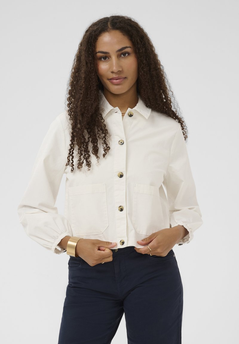 Chaqueta color crema con cierre de botones al frente, dos bolsillos en el pecho y puños fruncidos en las mangas. Combinada con pantalones azul marino, diseño minimalista.