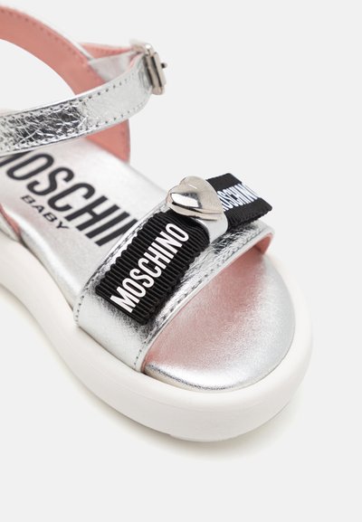 Sandale en argent métallique avec une large bride noire arborant le logo blanc "Moschino", un accent en forme de cœur argenté et une doublure intérieure rose. Semelle blanche.
