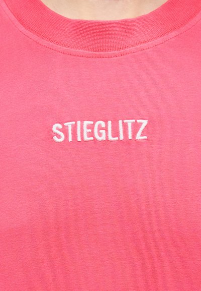 Κοντινό πλάνο ροζ μπλούζας με στρογγυλή λαιμόκοψη και λευκό κεντημένο κείμενο "STIEGLITZ" στο κέντρο της περιοχής του στήθους.
