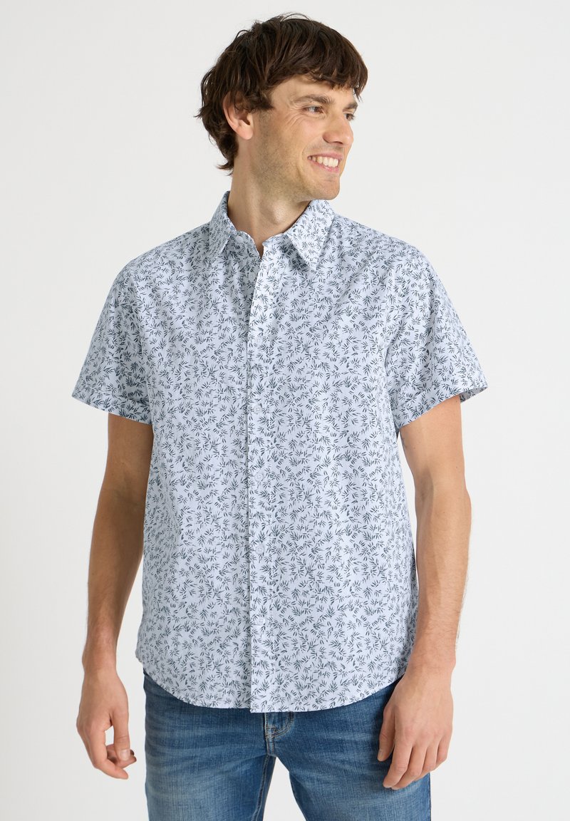 Jeune homme souriant, portant une chemise à manches courtes bleu clair avec un petit motif de feuilles foncées et un jean bleu, debout devant un fond blanc.