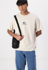 Beige cotton t-shirt with a central blue logo. Paired with light blue denim jeans and a small black crossbody bag with zippers.