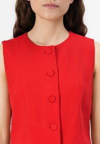 Vestido sin mangas rojo con cuello redondo y cinco botones circulares en la parte delantera. Fabricado en un tejido texturizado. Sin patrones ni acentos visibles.