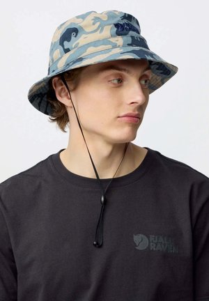 Jeune personne portant un chapeau bob camouflage bleu et beige avec une sangle sous le menton et un t-shirt noir Fjällräven, regardant vers la droite.