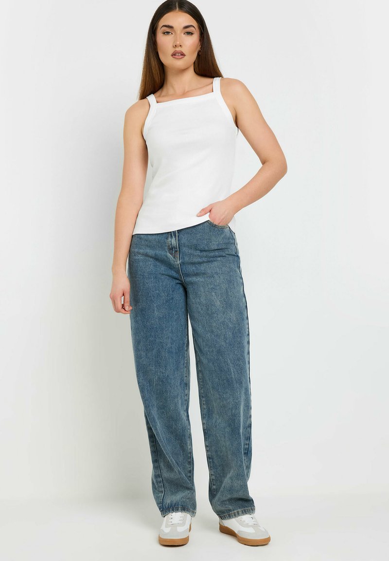 Top bianco senza maniche con scollatura quadrata, abbinato a jeans in denim azzurro chiaro a vestibilità morbida. Il modello indossa sneakers bianche con dettagli beige.