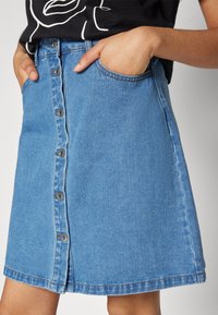 Lichtblauwe denim rok met A-lijn ontwerp, voorzijde met knoopsluiting en zijzakken. Heeft zichtbare stiksels en een zoomlijn boven de knie.