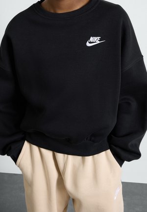 Henkilö, joka pitää yllään mustaa Nike-hupparia ja beigen värisiä Nike-treenihousuja, kädet taskuissa seisoen yksinkertaista taustaa vasten.