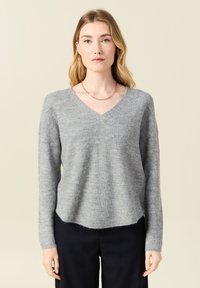 Grauer, gestrickter Pullover mit V-Ausschnitt, langen Ärmeln und lockerer Passform. Weicher Stoff und leicht fransiger Saum. Getragen mit dunklen Hosen.