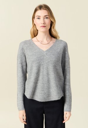 Suéter gris de punto con escote en V, mangas largas y corte holgado. Textura suave y detalle de dobladillo ligeramente deshilachado. Llevado con pantalones oscuros.