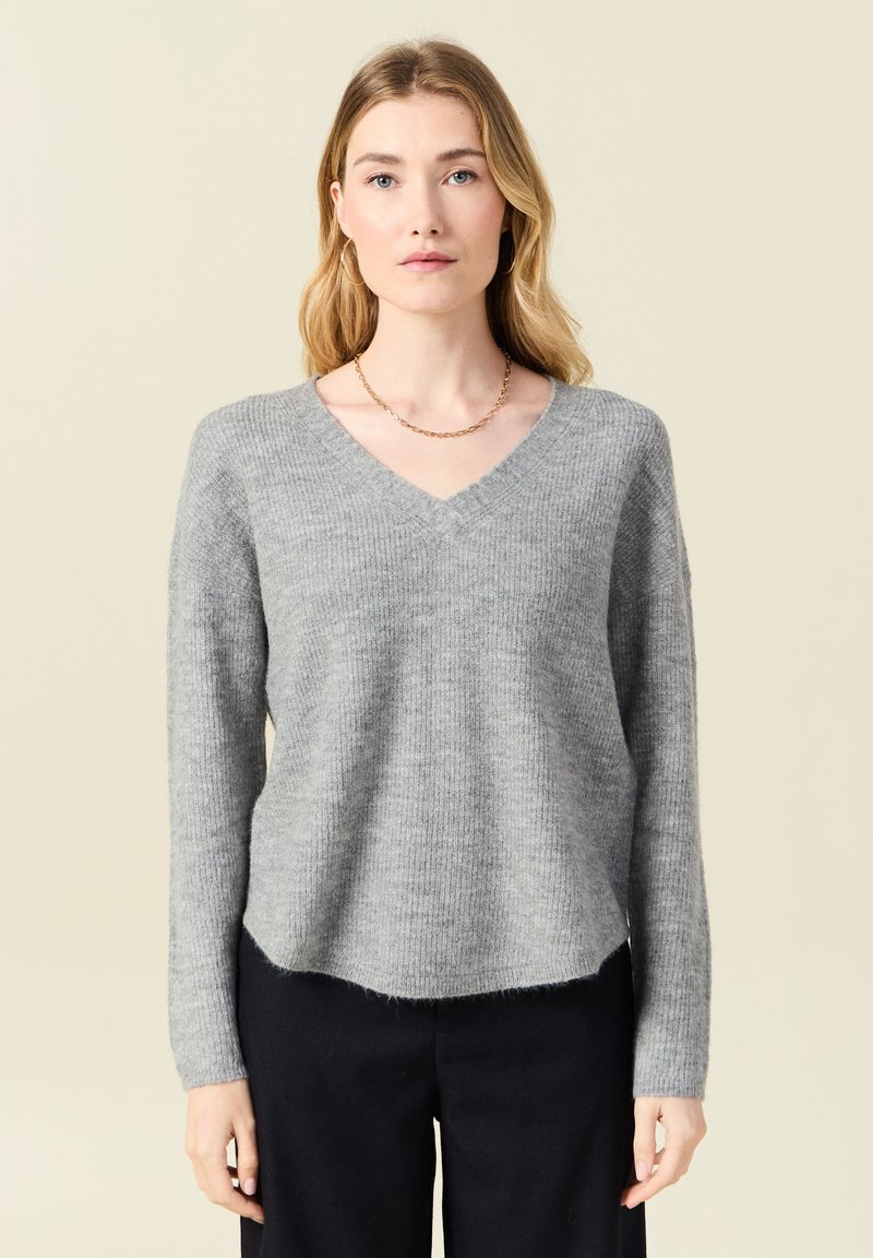 Grauer, gestrickter Pullover mit V-Ausschnitt, langen Ärmeln und lockerer Passform. Weicher Stoff und leicht fransiger Saum. Getragen mit dunklen Hosen.