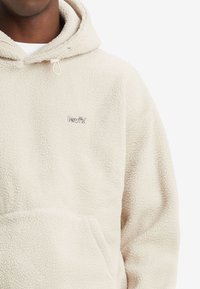 Beżowa polarowa bluza z kapturem, z kieszenią typu kangur, elastycznym sznurkiem w kapturze oraz haftowanym logo "Levi's" na piersi. Miękka, teksturowana tkanina.