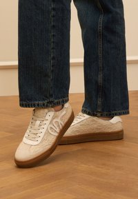 Lace-up Sneaker in Creme mit weißen Akzenten und einer braunen Gummisohle, die ein strukturiertes Muster aufweisen. Getragen mit dunkelblauen Jeans.