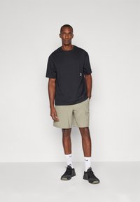 Zwarte T-shirt met korte mouwen en een klein patch, gecombineerd met licht khaki shorts. Zwarte sportschoenen en witte sokken maken de outfit compleet.